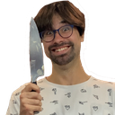 A man with glasses and a beard holds a kitchen knife while wearing a wide, chaotic, and mischievous grin.