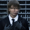A man in a black suit and tie with black gloves stares intensely with a digitally distorted, stretched facial expression.