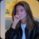 A woman with brown hair wearing a black leather jacket covering her mouth with her hand while laughing.