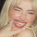 Blonde woman winking and smiling with her hand near her face in a playful gesture.