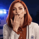 A woman with red hair wearing a white medical coat blows a kiss toward the camera with blue emergency lights in the backgrou…