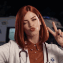 A woman with red hair wearing a lab coat and stethoscope winks and makes a peace sign in front of an ambulance.