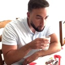 A man with a beard looking down while holding a glass of liquid to his lips.