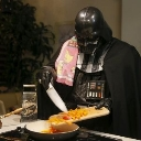 Darth Vader wearing a pink patterned apron while carefully chopping vegetables on a cutting board in a kitchen.