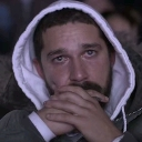 Shia LaBeouf wearing a white-lined hoodie with his hands clasped in front of his face, looking intensely at the camera.