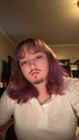 A person with purple hair and a drawn-on mustache looking directly at the camera with a deadpan expression.