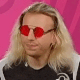A person with long blonde hair wearing bright red sunglasses looking directly at the camera with a serious, stoic expression.