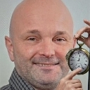 A bald man smiling and pointing his finger at a silver pocket watch.