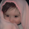 A baby with brown hair peeking out from under a soft pink blanket with a shy expression.