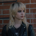 Young woman with blonde shaggy hair and a leather jacket looking to the side with a skeptical expression.