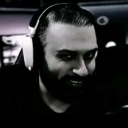 A bearded man wearing headphones looking at the camera with a dark, suspicious smirk.