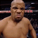 A fighter with a bald head makes an intense, angry, and menacing facial expression while looking directly at the camera.