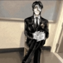 Man in a black suit sitting with hands clasped and a serious, suspicious expression.