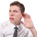 A young man in a white shirt and tie cups his hand behind his ear with a confused expression.