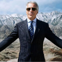 Man in a dark suit and sunglasses standing with arms spread wide in front of snow-capped mountains.