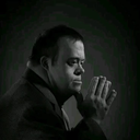 A man in a dark suit poses with his hand near his chin while looking thoughtfully to the side.