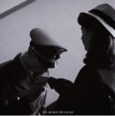 A soldier in uniform stands still while a woman in a hat pins a medal onto his chest in a black and white scene.