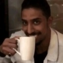 A man with a mischievous smirk holds a white mug to his mouth.