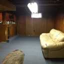 Basement interior featuring wood-paneled walls, a beige sofa, and bright overhead fluorescent lighting.