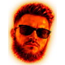 Man with a beard and dark sunglasses looking directly forward under a warm orange light.