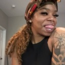 A woman with a curly ponytail and arm tattoo sticks her tongue out playfully.