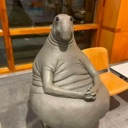 Wosh sculpture sitting with hands folded in its lap with a neutral, seal-like face.