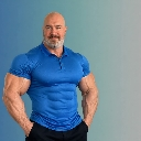 Muscular man with a beard stands with hands in his pockets wearing a tight blue polo shirt.