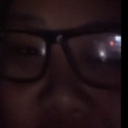 A person wearing black-rimmed glasses stares intensely from the shadows.