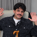 A man with curly hair and a mustache waving both hands with a cane visible at the bottom.