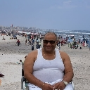 A man wearing sunglasses and a white tank top sits in a wheelchair on a sunny, crowded beach.