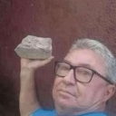 An older man with glasses holds a rock up near his head with a serious, blank expression.
