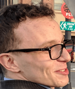 A man with glasses gives a tight-lipped, slightly awkward smile.