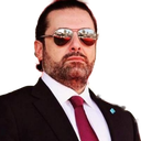 A man wearing a black suit, white shirt, red tie, and dark sunglasses looking directly at the camera with a serious expres...